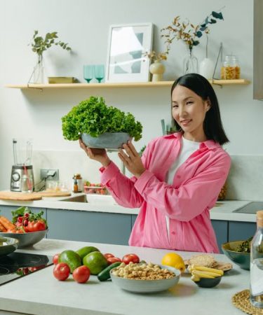 Woman in a kitchen holding fresh lettuce, surrounded by healthy ingredients like tomatoes and avocados, promoting healthy eating.